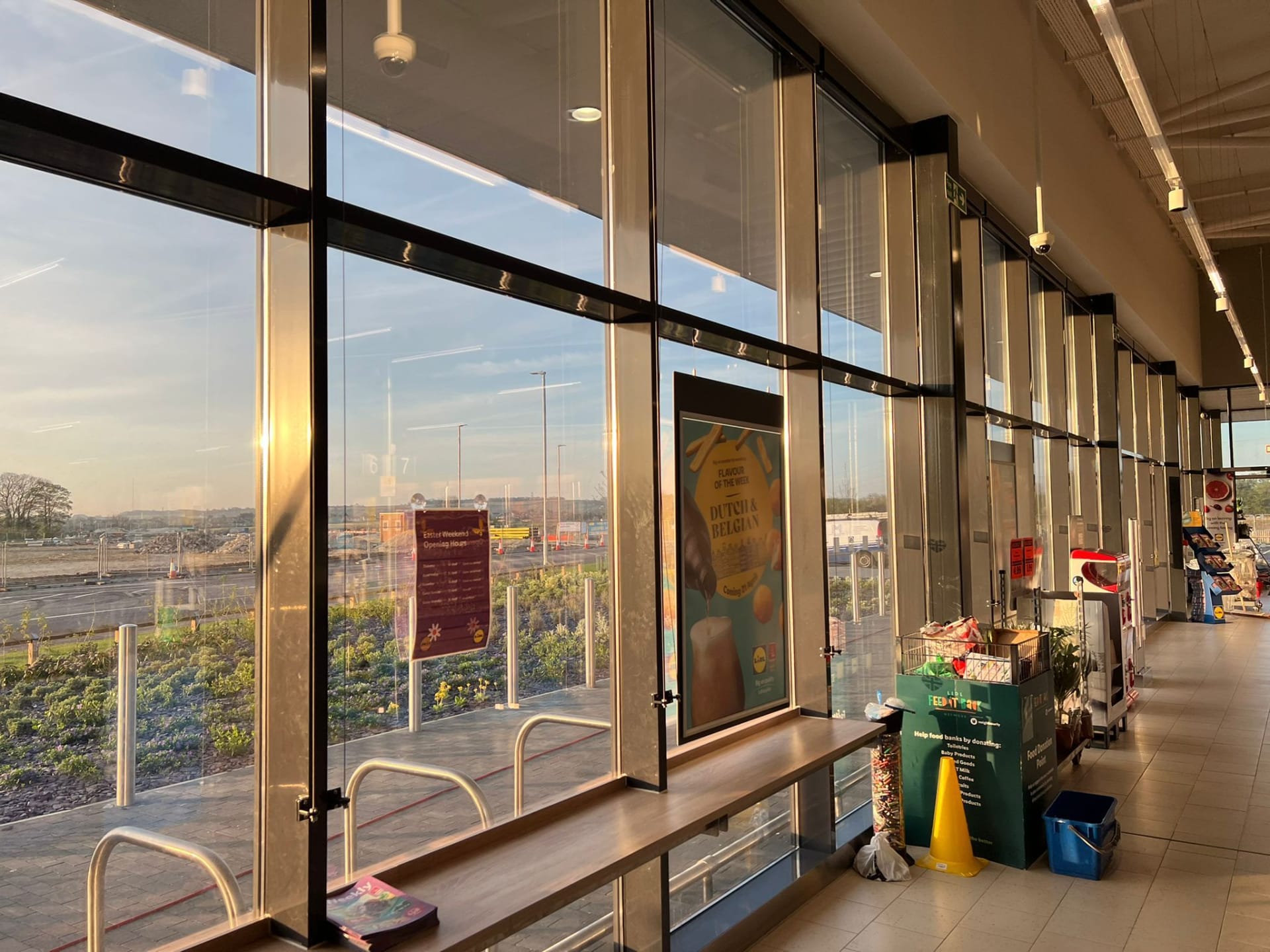 Supermarket interior with large windows bathed in golden sunlight, looking out onto a construction site. Inside, a wooden counter, promotional posters, and a food bank donation point are visible.