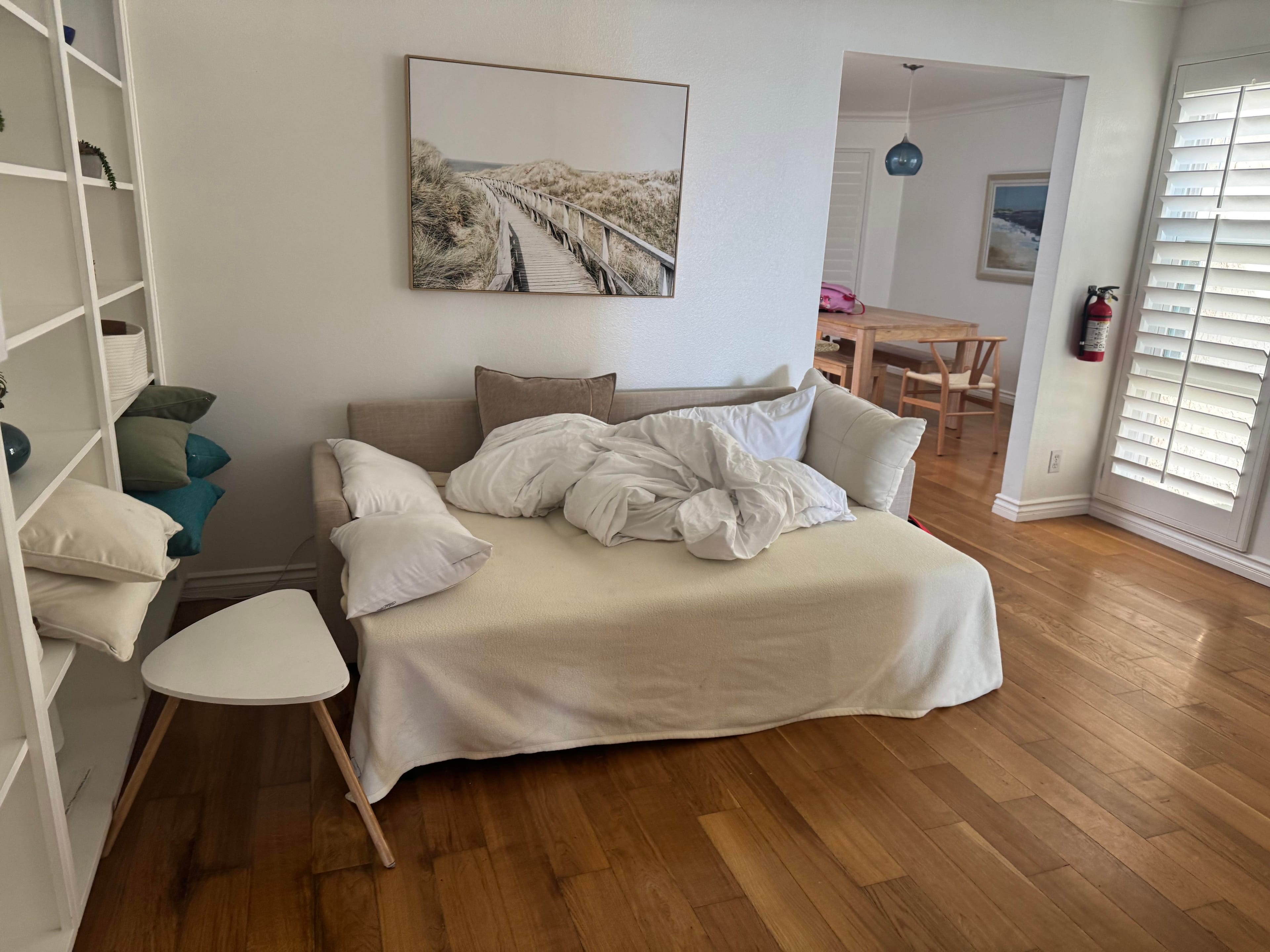 A bright, minimalist living room features a daybed with rumpled white bedding, a white shelving unit, and hardwood floors. An artwork of a beach boardwalk hangs above the daybed, and a dining room is visible through an archway.