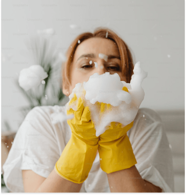 Person in yellow gloves playfully holding a large amount of white soap suds, with bubbles splashing around their face.