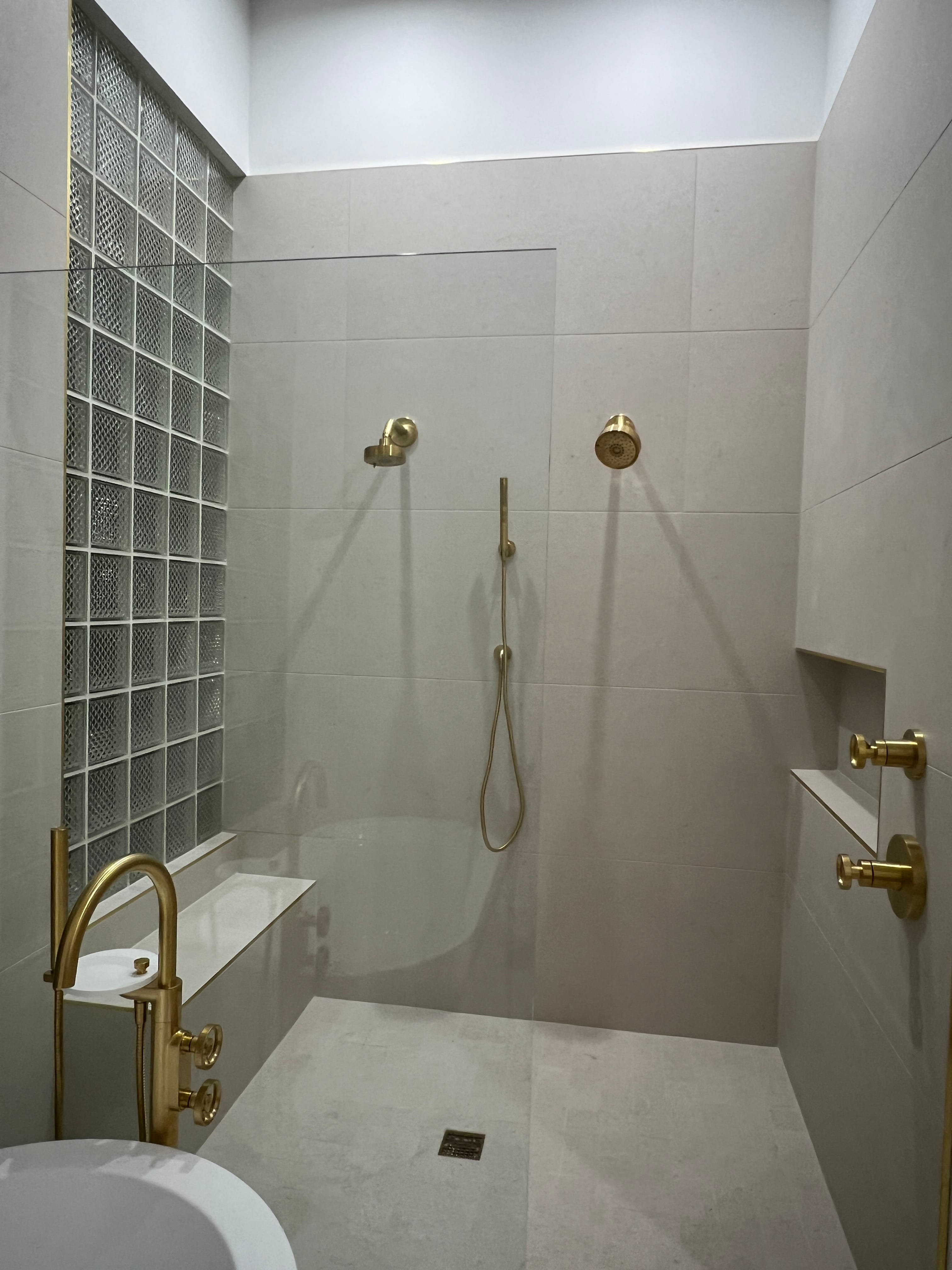 Modern bathroom featuring a walk-in shower with gold fixtures, beige tiles, a glass block wall, and a freestanding tub.