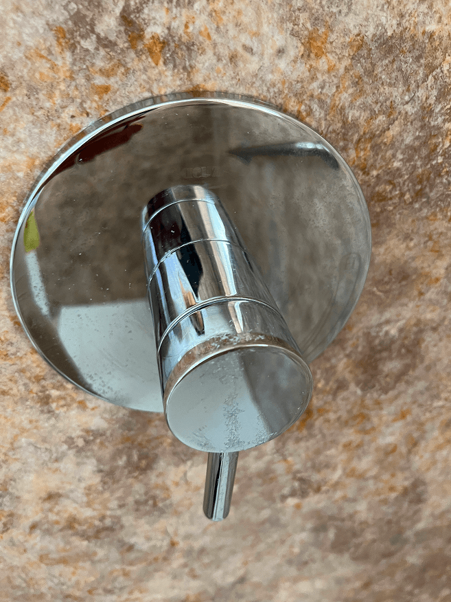 Chrome shower control with a single lever handle and round plate, faintly branded 'TOTO', on a textured brown wall with visible water spots.