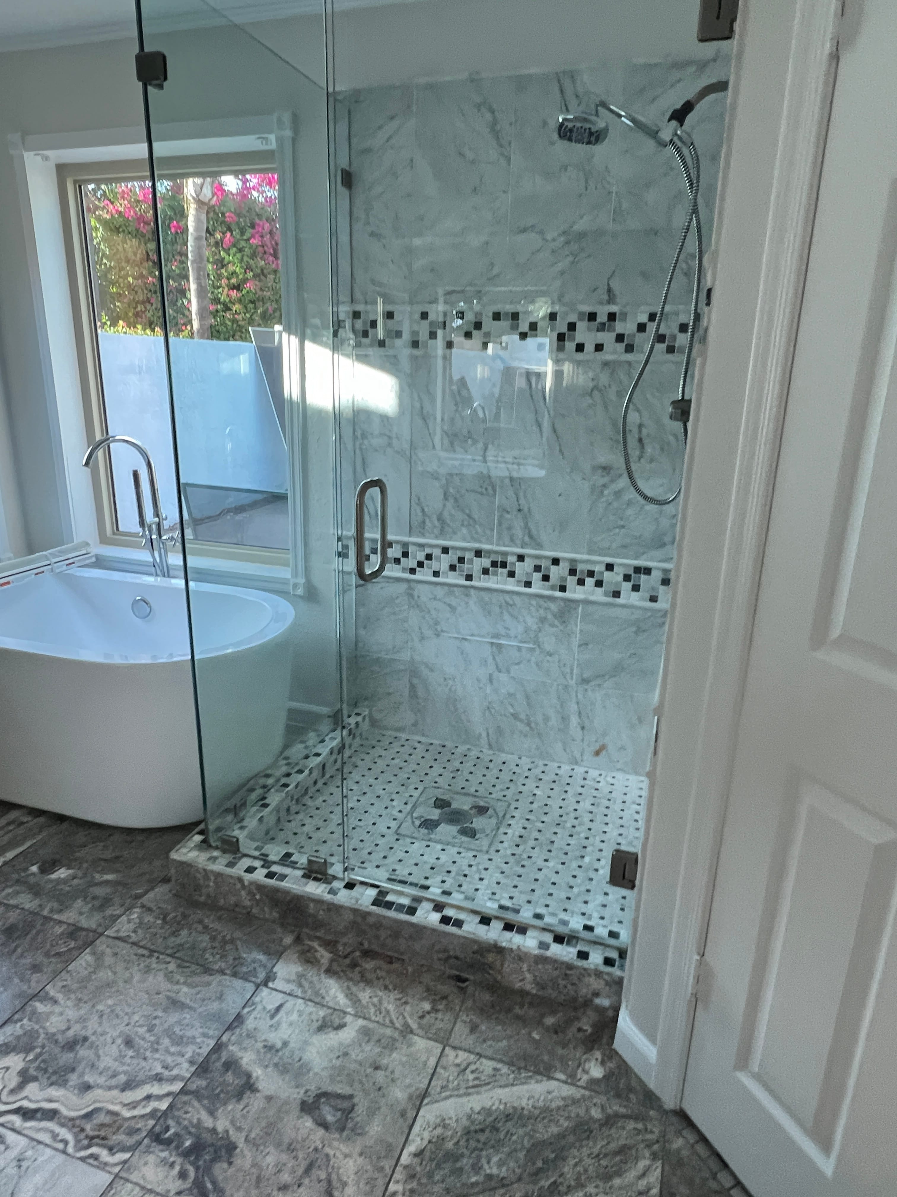 A modern bathroom with a white freestanding tub by a window and a glass shower stall featuring marble-look tiles and a black and white mosaic accent. The floor is tiled in grey.