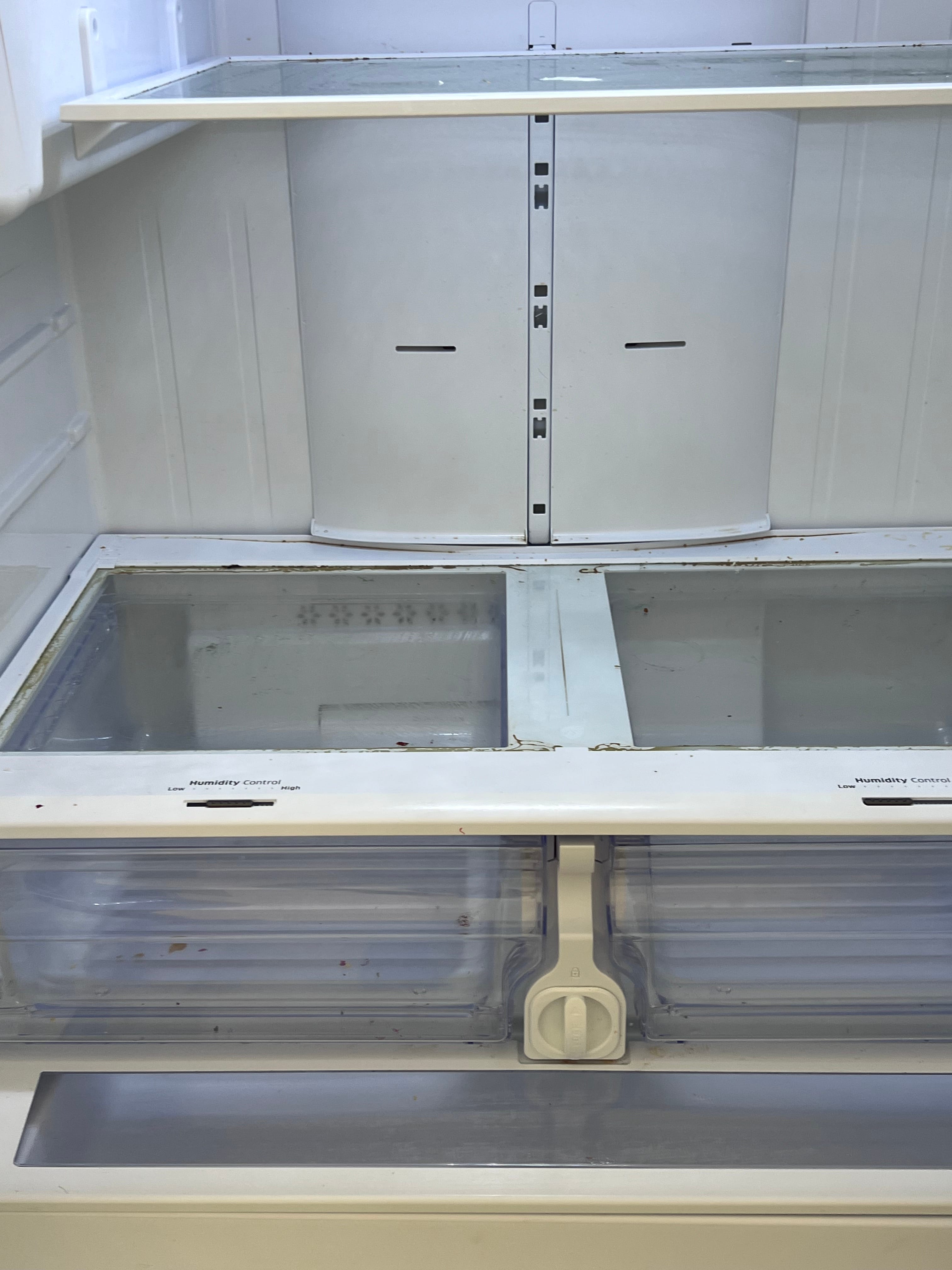Inside an empty, dirty refrigerator, showing visible grime on glass shelves, white plastic walls, and clear crisper drawers with 'Humidity Control' labels.