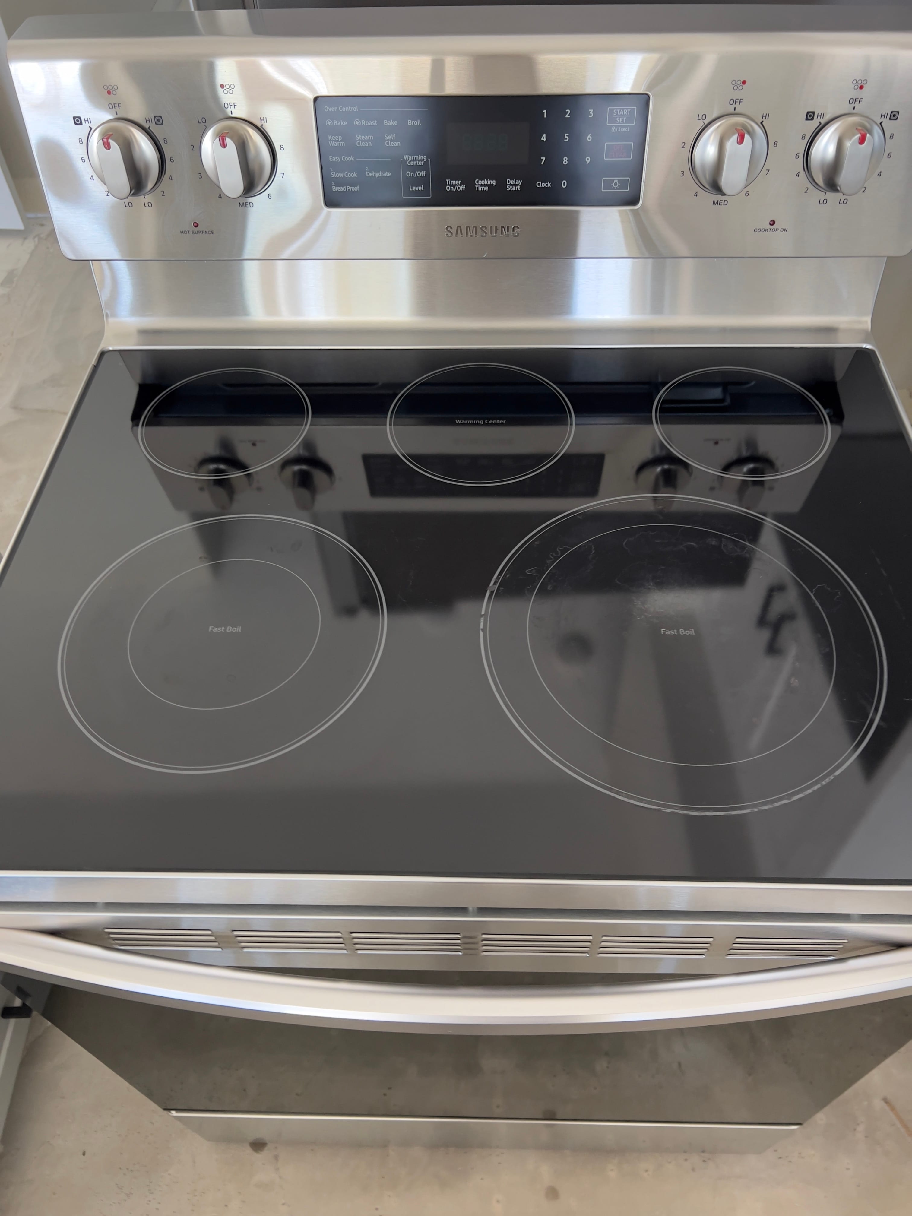 Clean, stainless steel Samsung electric range with a black glass cooktop showing four burners, two with 'Fast Boil,' and a central warming center. The front panel has five burner control knobs and a digital oven control panel for functions like Bake, Roast, and Self Clean.