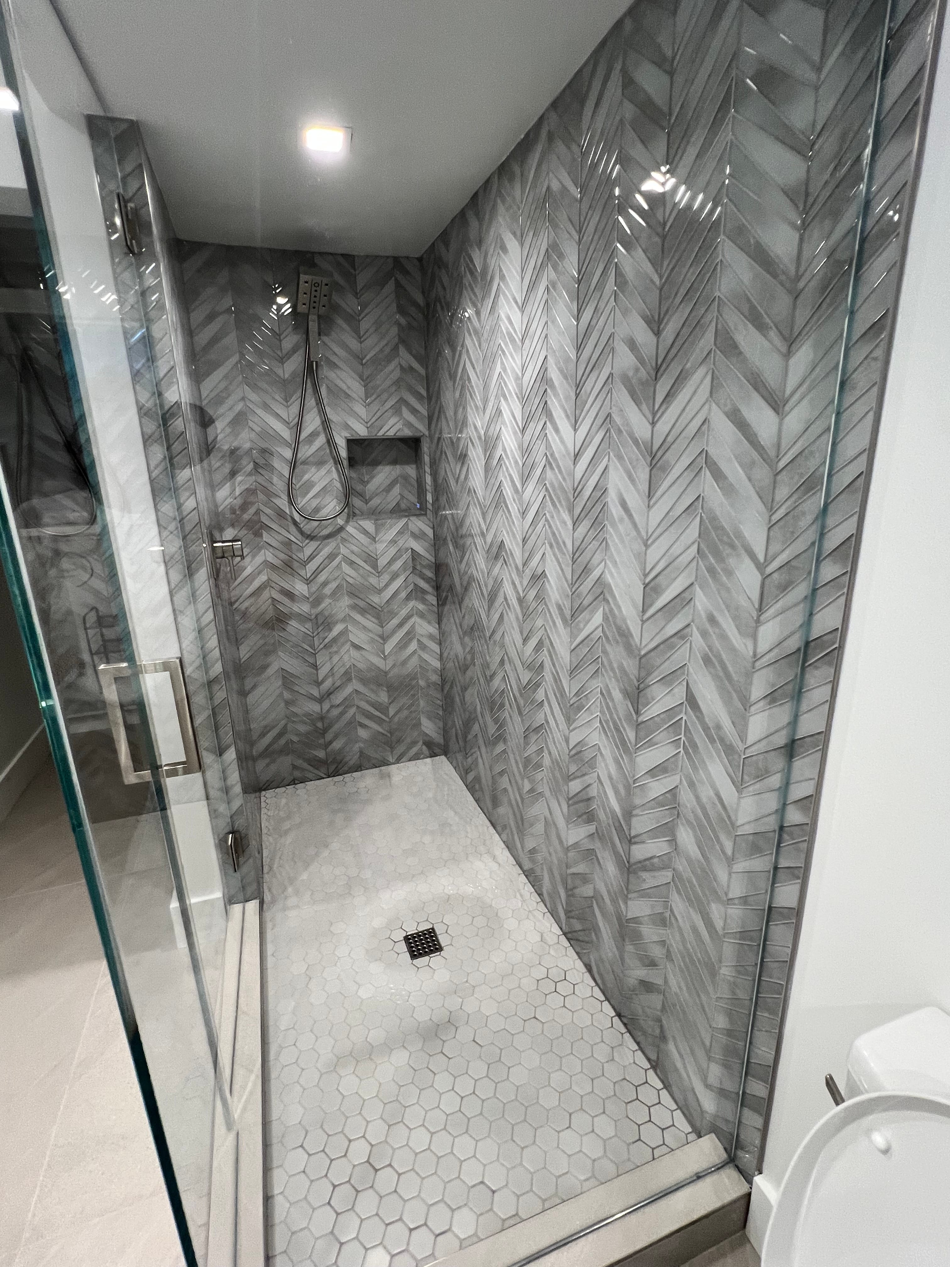 New, modern walk-in shower featuring grey chevron tiled walls, a white hexagonal floor, and a glass enclosure.