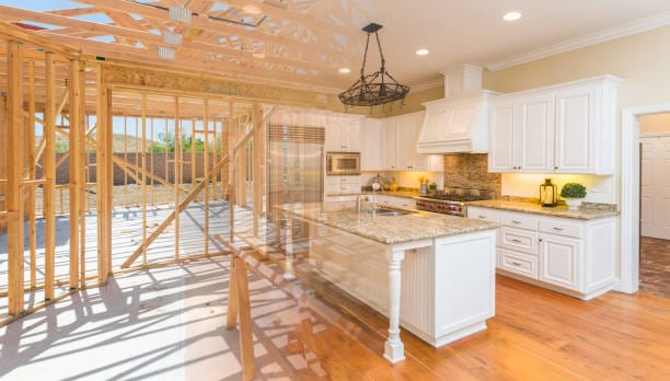 A split image of home construction progress: unfinished wooden framing on the left blends into a luxurious, complete white kitchen with granite countertops, stainless steel appliances, and a large island on the right.
