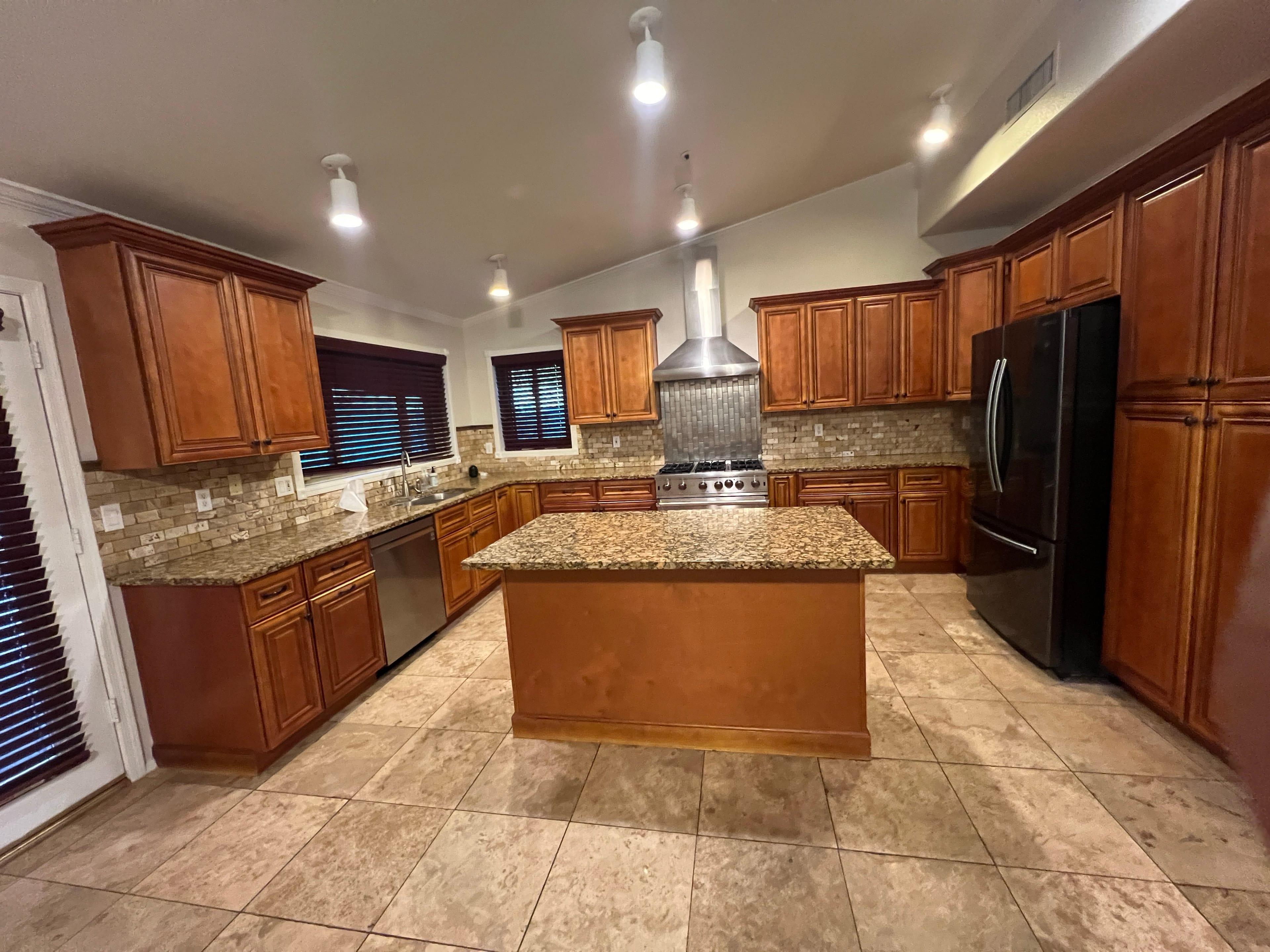 Clean kitchen with brown wooden cabinets, granite countertops, stone backsplash, stainless steel appliances, a large island, and light tile flooring.