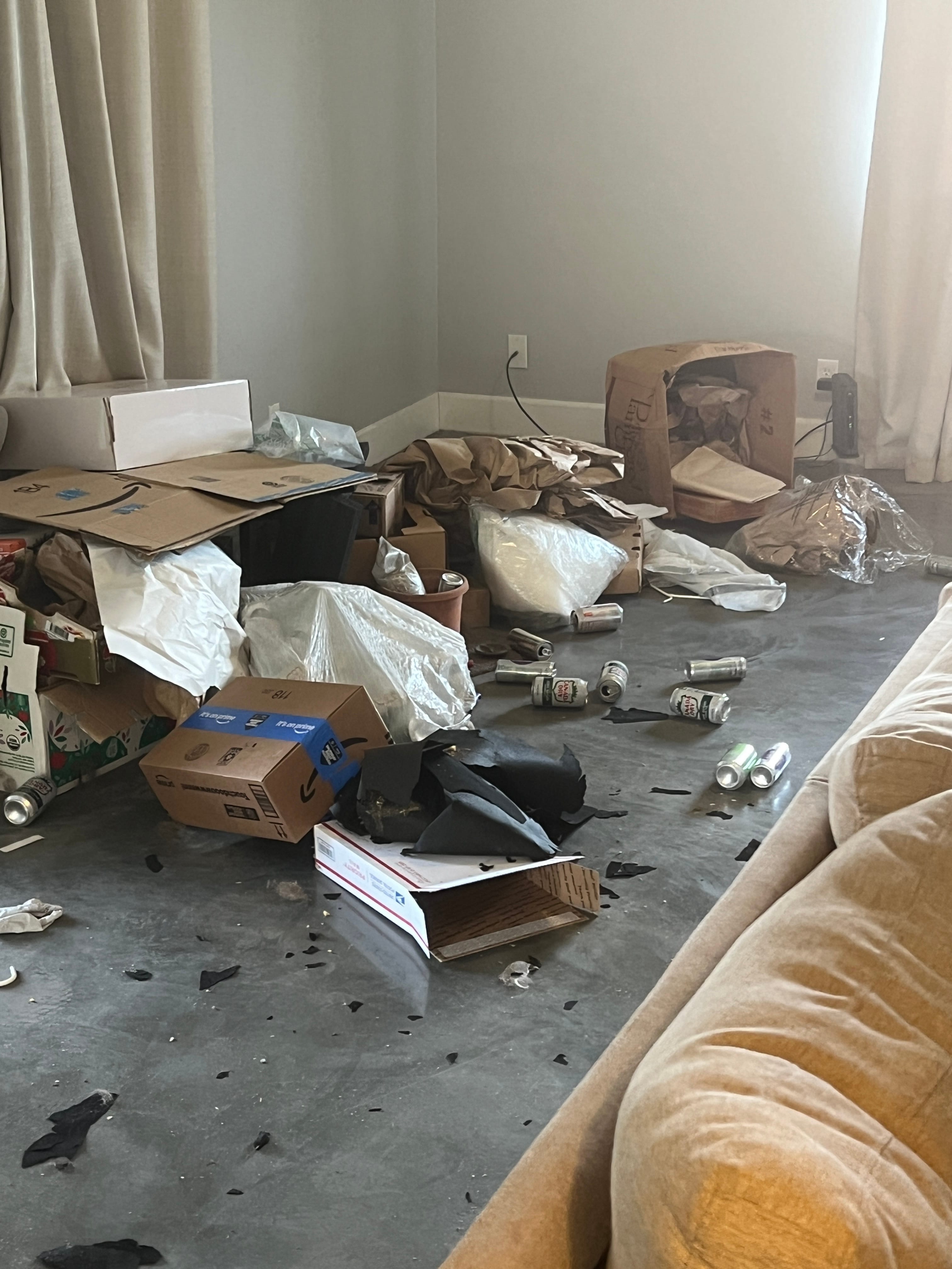 A very messy living room filled with cardboard boxes, crumpled packaging, plastic bags, empty soda cans, and torn black material scattered on a dark floor, with a light sofa on the right.