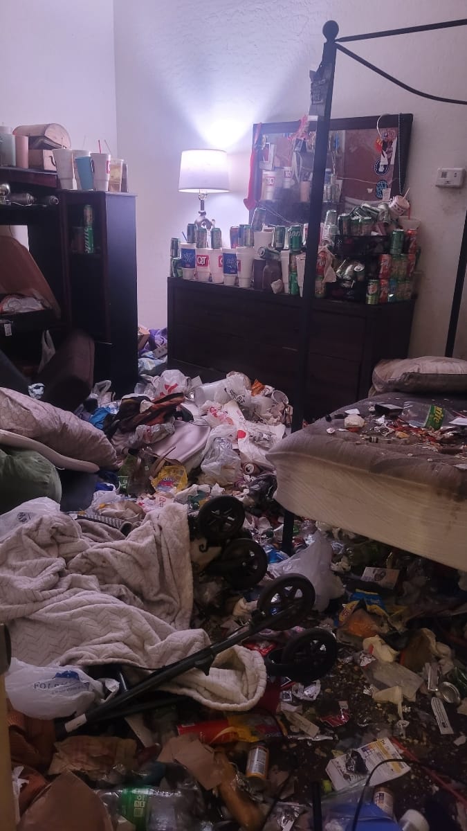 A very messy room filled with trash, empty cups, and soda cans covering the floor, bed, and furniture, suggesting a severe hoarding situation.