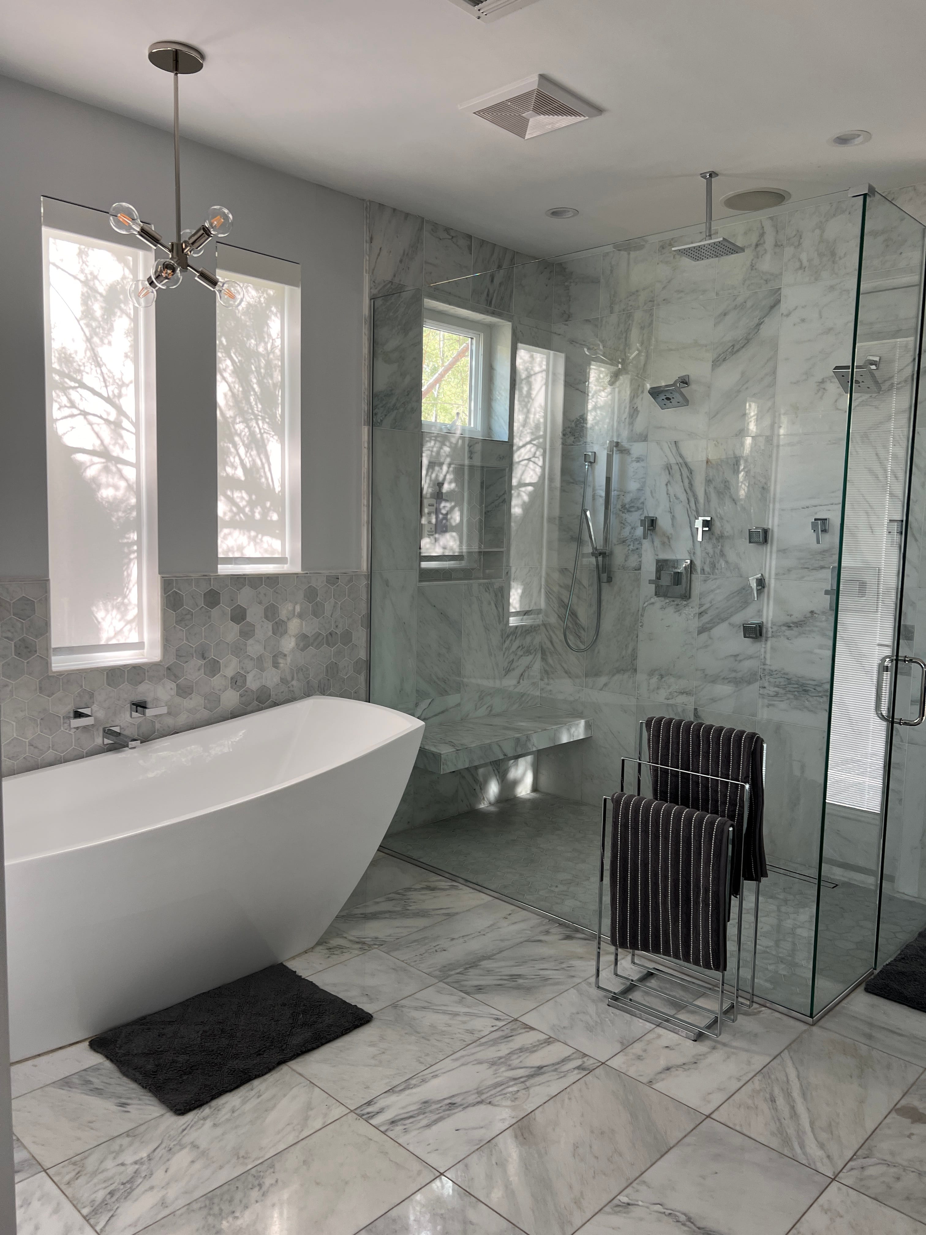 Bright, modern bathroom with a white freestanding tub, walk-in shower featuring light grey marble walls and bench, hexagonal accent tiles, and chrome fixtures.