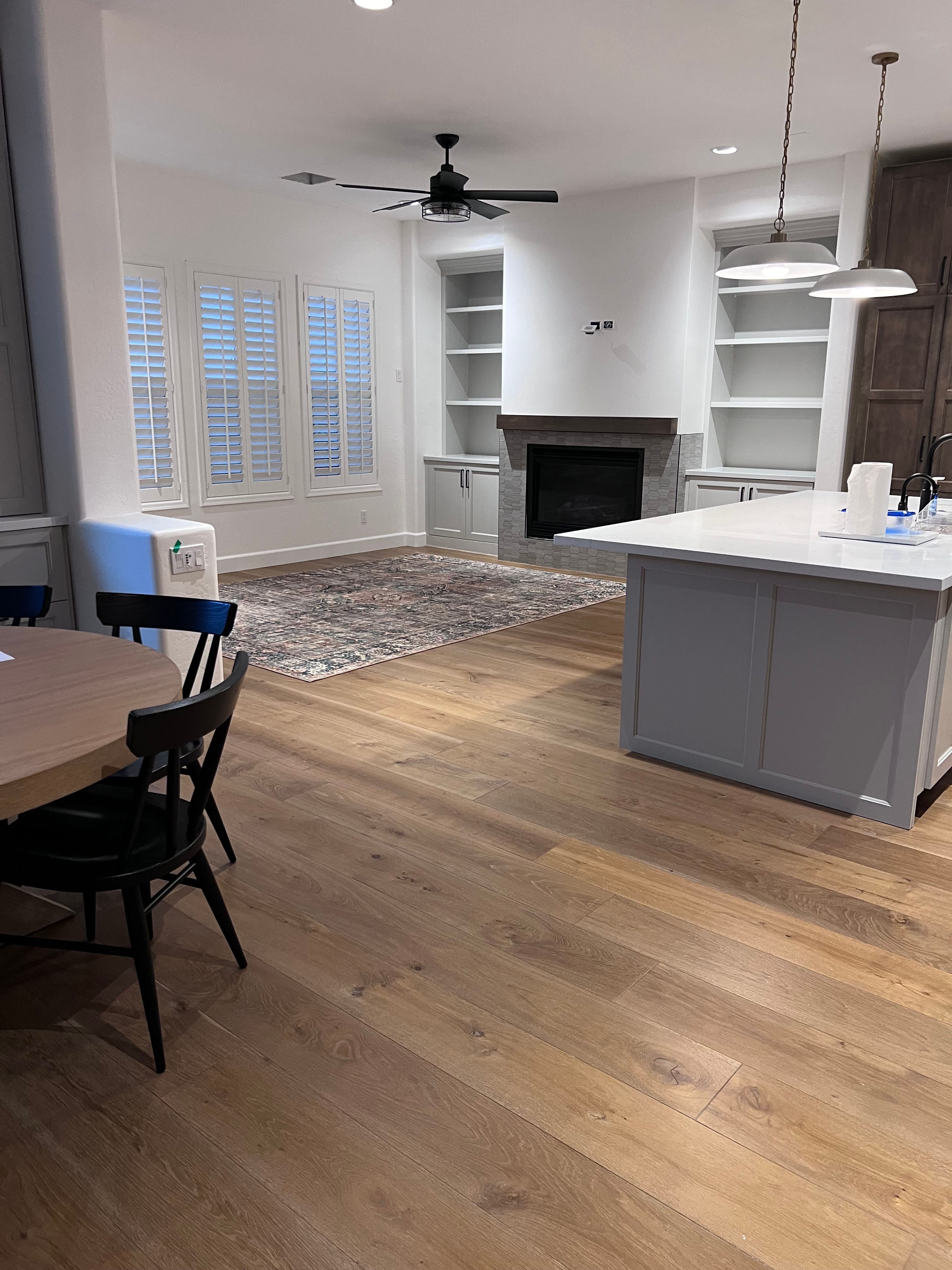 A modern open concept living and dining area featuring light hardwood floors, a patterned area rug, a fireplace with built-in shelves, a kitchen island with pendant lights, and a dining table.