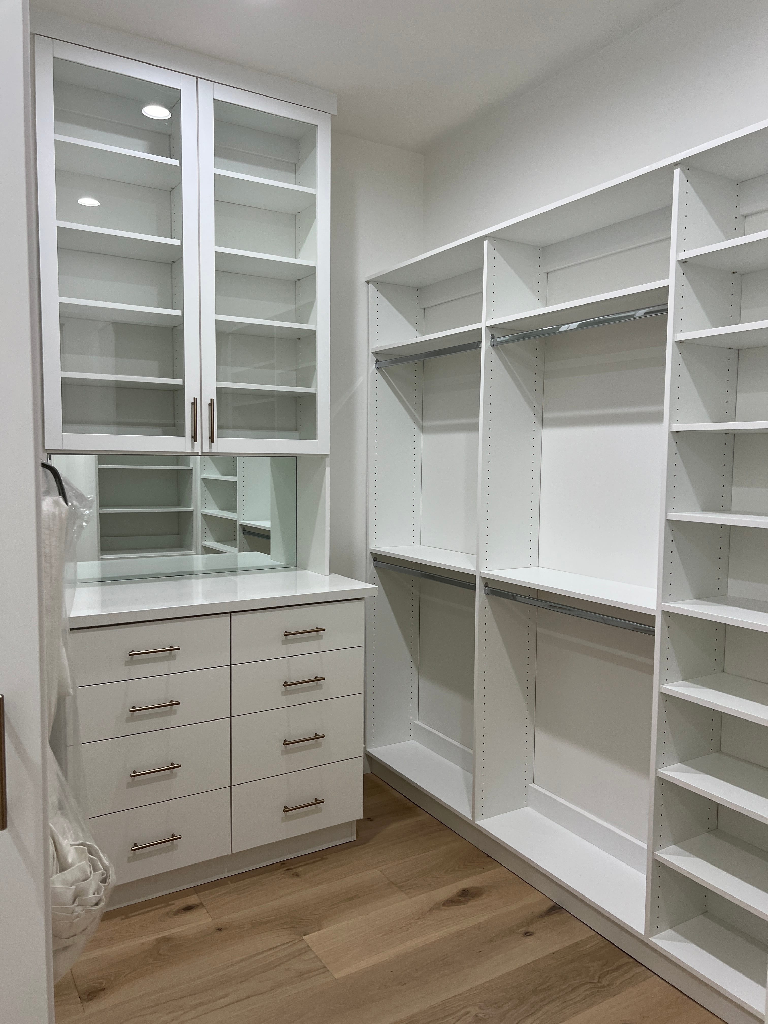 A bright, white custom walk-in closet with glass-front cabinets, internal lights, a mirrored counter over drawers with bronze handles, open shelving, and hanging rods, all on light wood flooring.