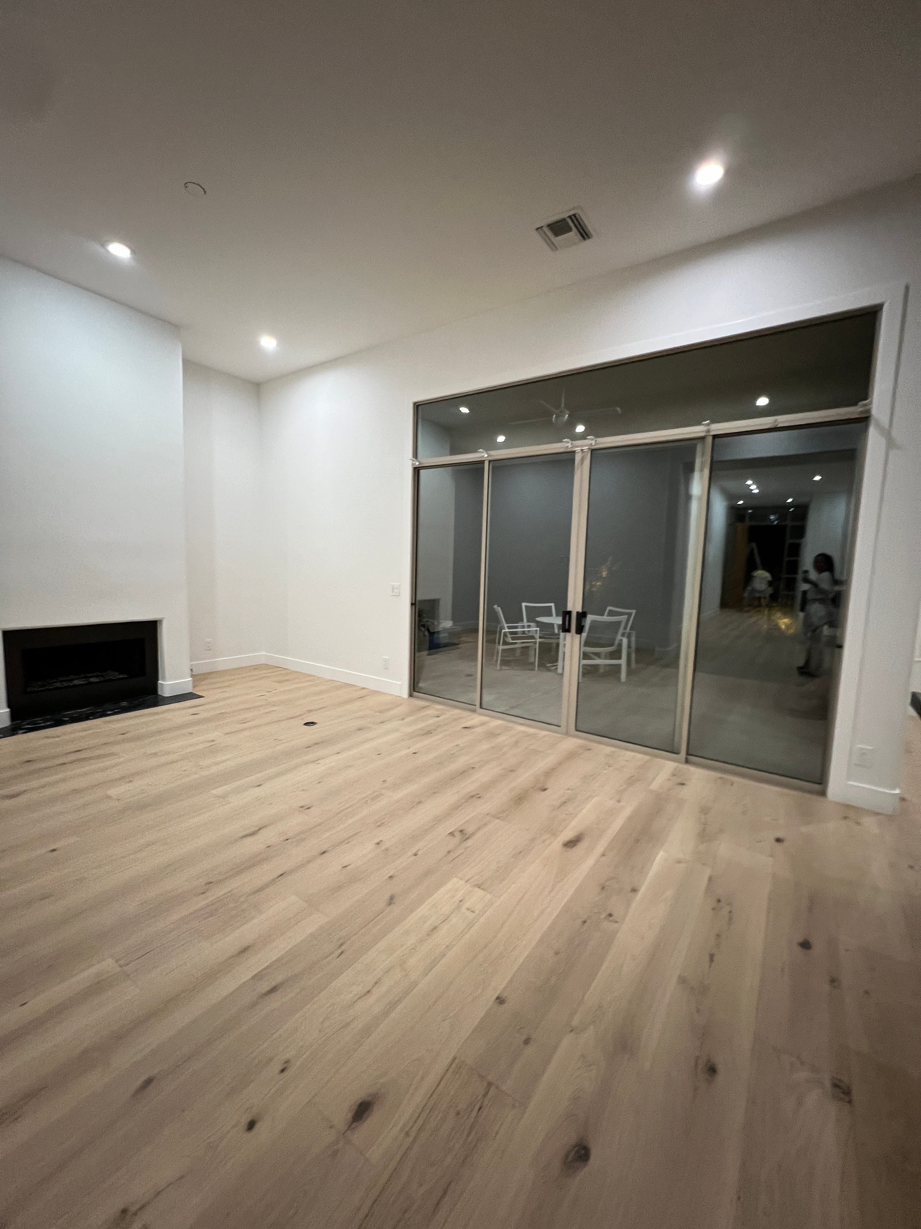 Empty, newly built living room with light wooden floors, white walls, a modern fireplace, and large glass sliding doors reflecting an adjacent area.