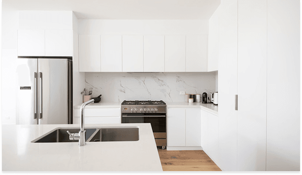 Bright, modern white kitchen featuring a stainless steel refrigerator, island with sink, marble backsplash, and a range oven, with warm wooden flooring.
