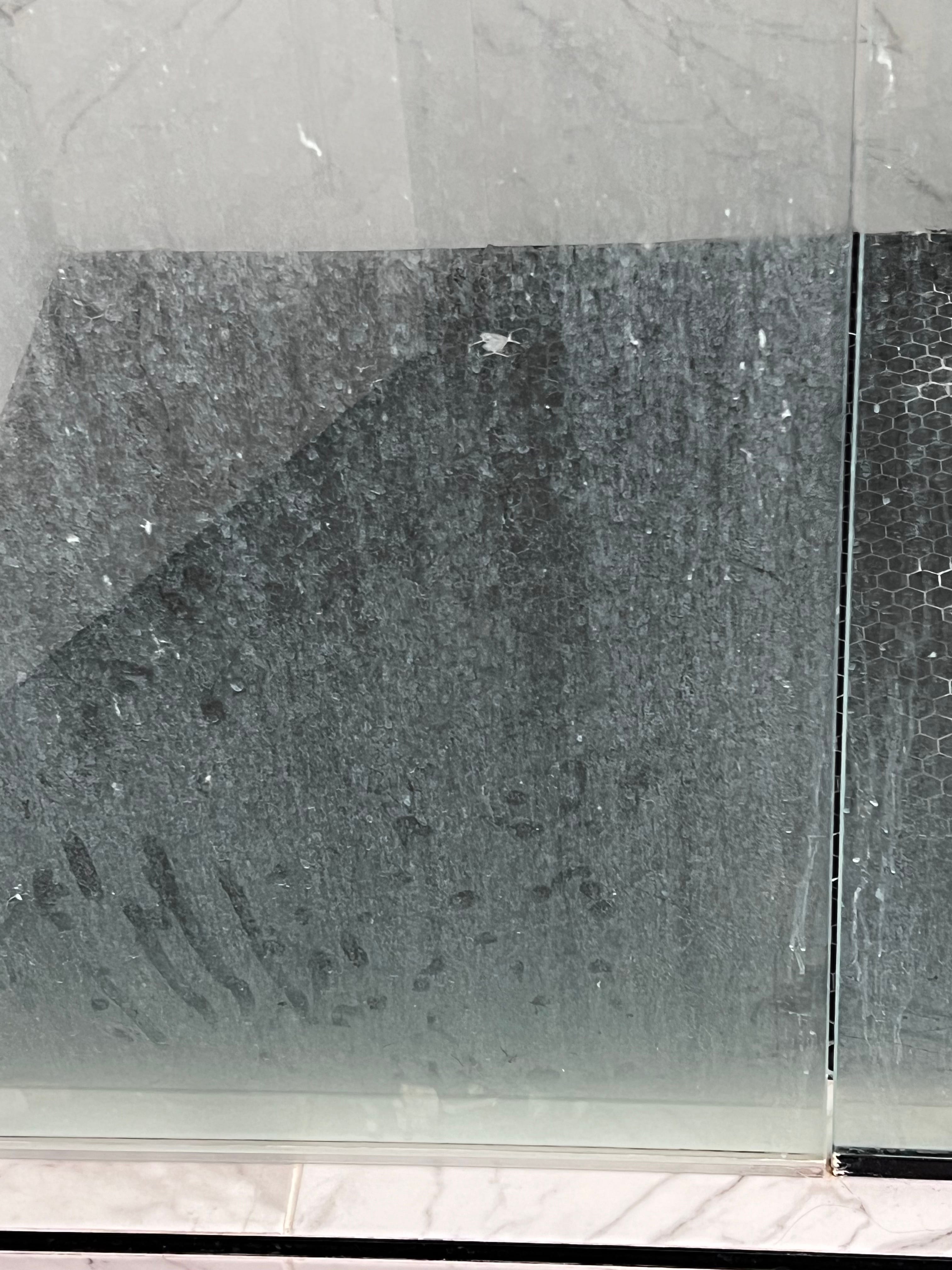 A dirty shower enclosure featuring dark textured walls and honeycomb tiles seen through a water-stained glass partition above a marble-patterned base.