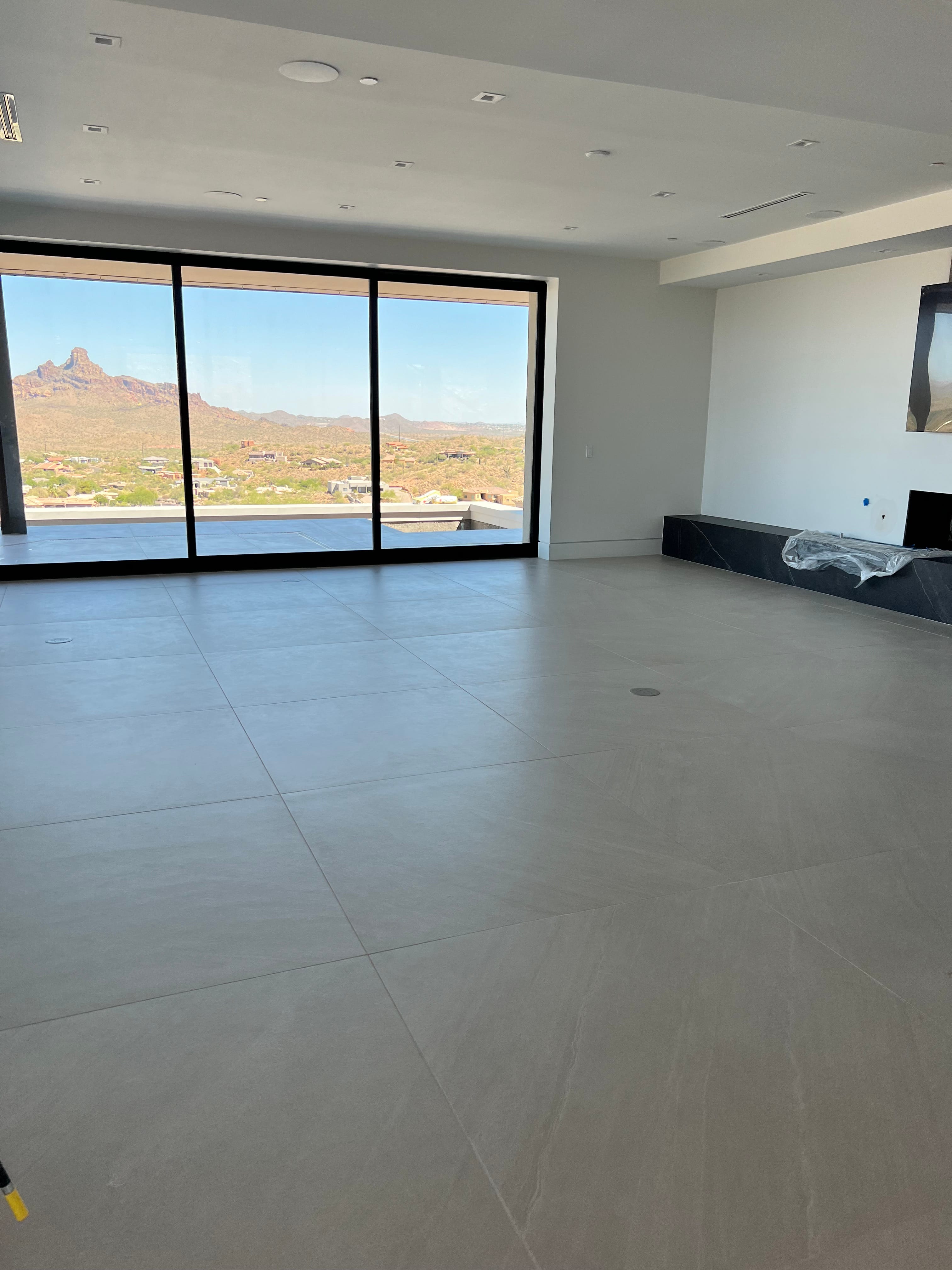 An empty, modern living room with light gray tile floors, recessed ceiling lights, and expansive windows offering a view of a desert mountain landscape with houses.