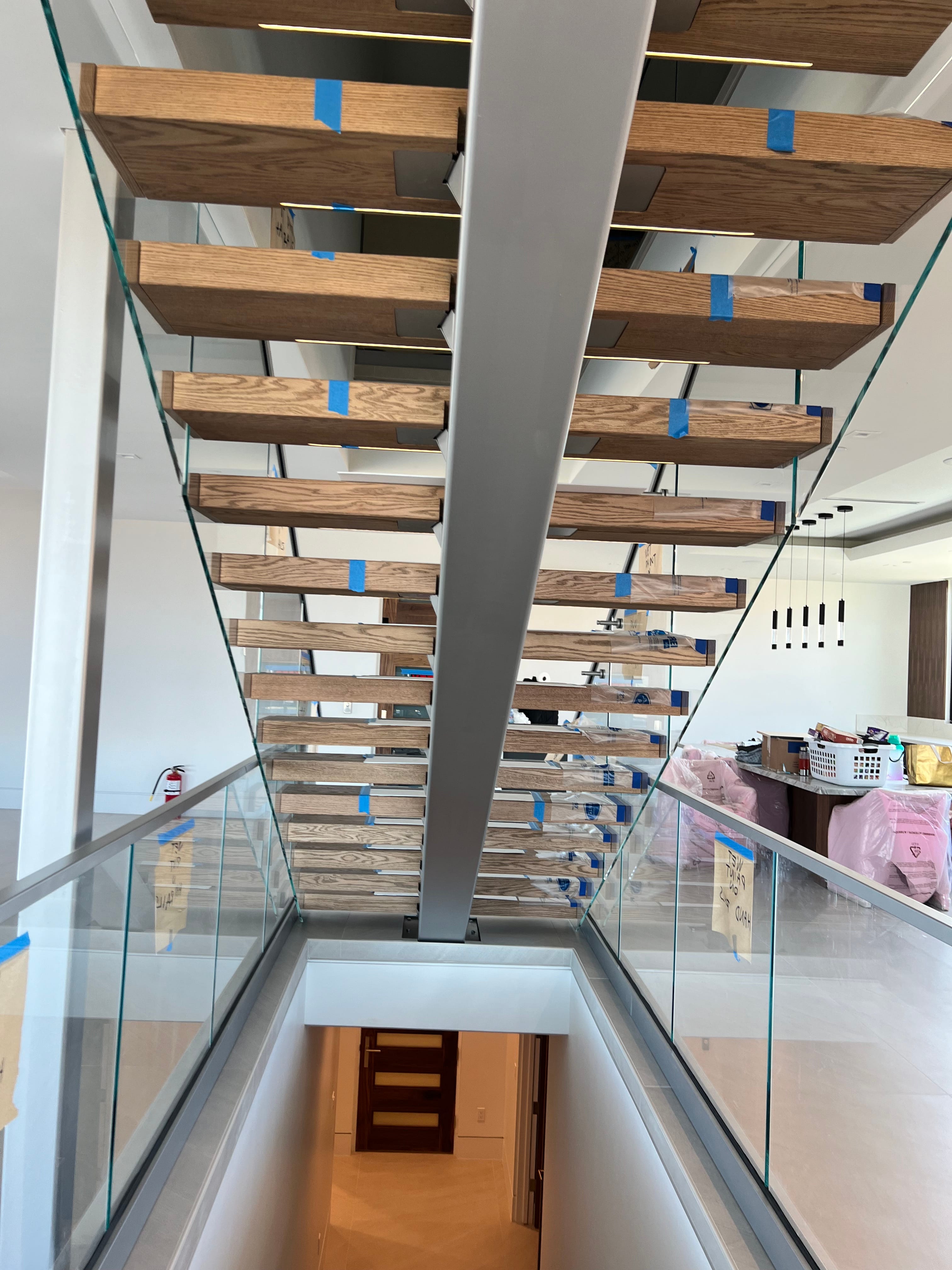 A modern floating staircase with light wood treads and clear glass railings, supported by a central metal beam, showing blue painter's tape from recent construction.