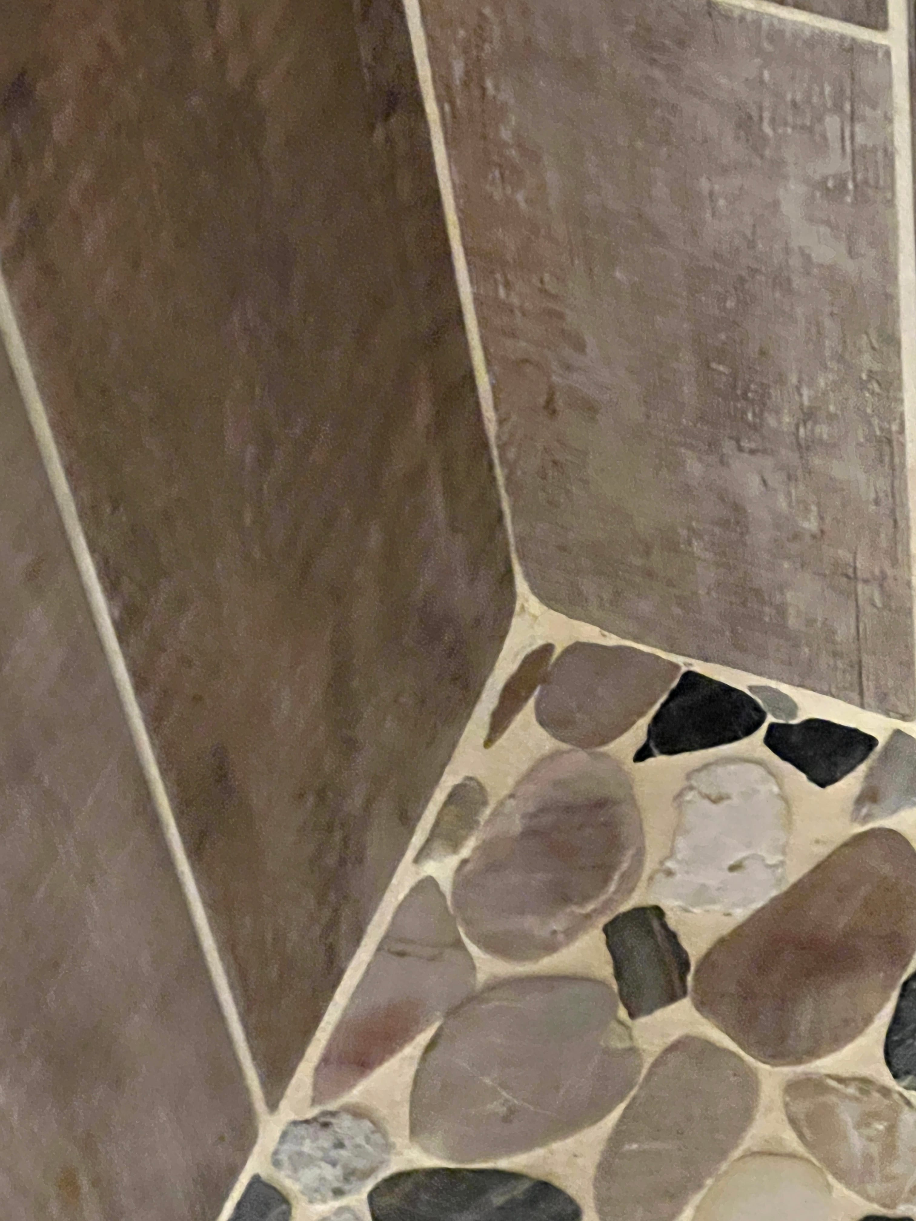 Close-up of a shower floor or wall, showing a corner where large, rectangular brown tiles meet a mosaic of smooth river pebbles in various natural colors, separated by light grout lines.