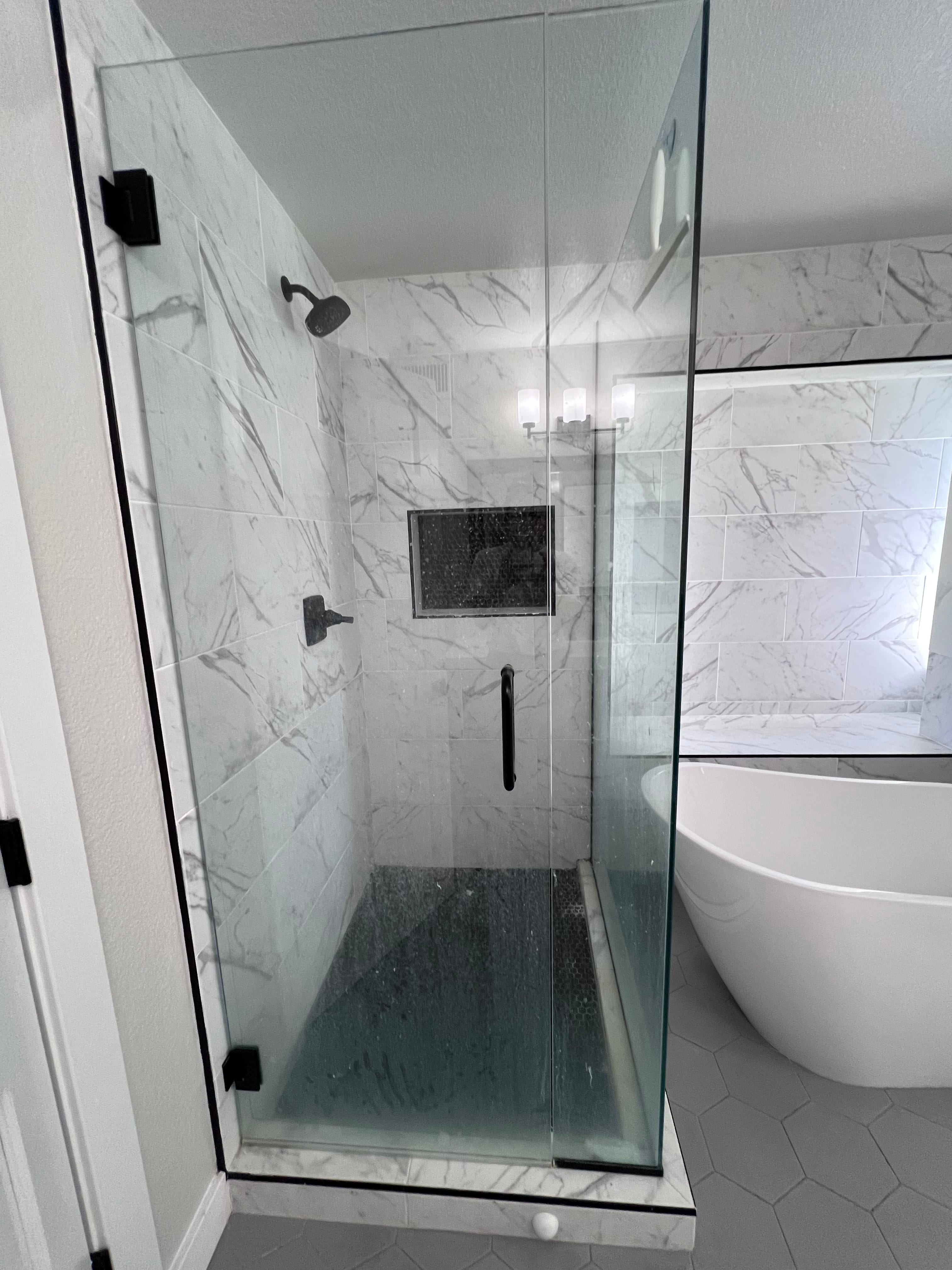 Modern bathroom featuring a glass shower stall with a wet and stained dark floor, white marble-look walls, black fixtures, and a freestanding white bathtub.